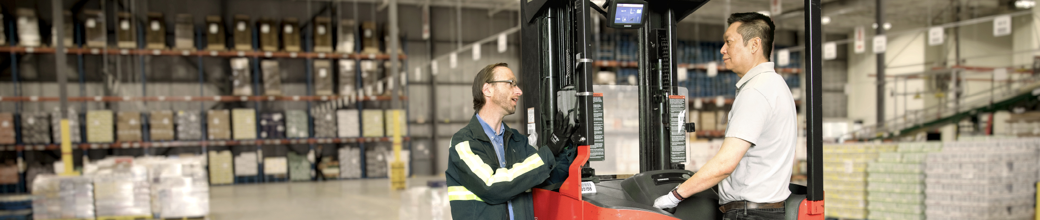 Deux employés de l’entrepôt de la LCBO en train de discuter. L’un des hommes se tient debout sur un chariot élévateur rouge dans l’entrepôt.