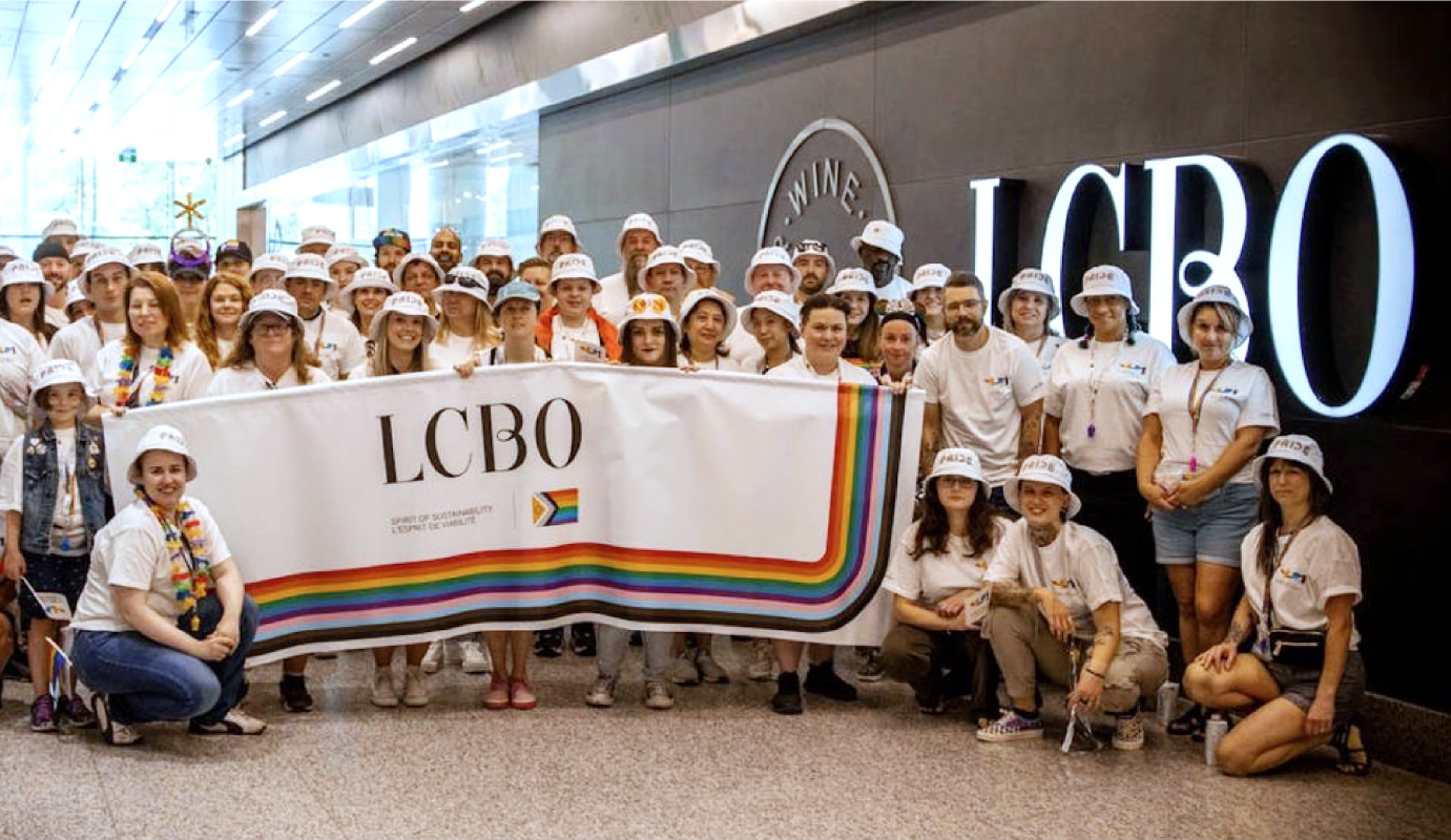 Cinquante personnes debout à côté d’une grande enseigne de la LCBO, tenant une bannière de la Fierté LCBO qui sera utilisée lors du défilé de la Fierté.