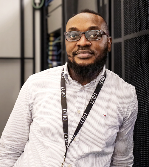 Un employé à lunettes, barbu et vêtu d’une chemise blanche se tient devant le bureau du service technologie au siège social de la LCBO.